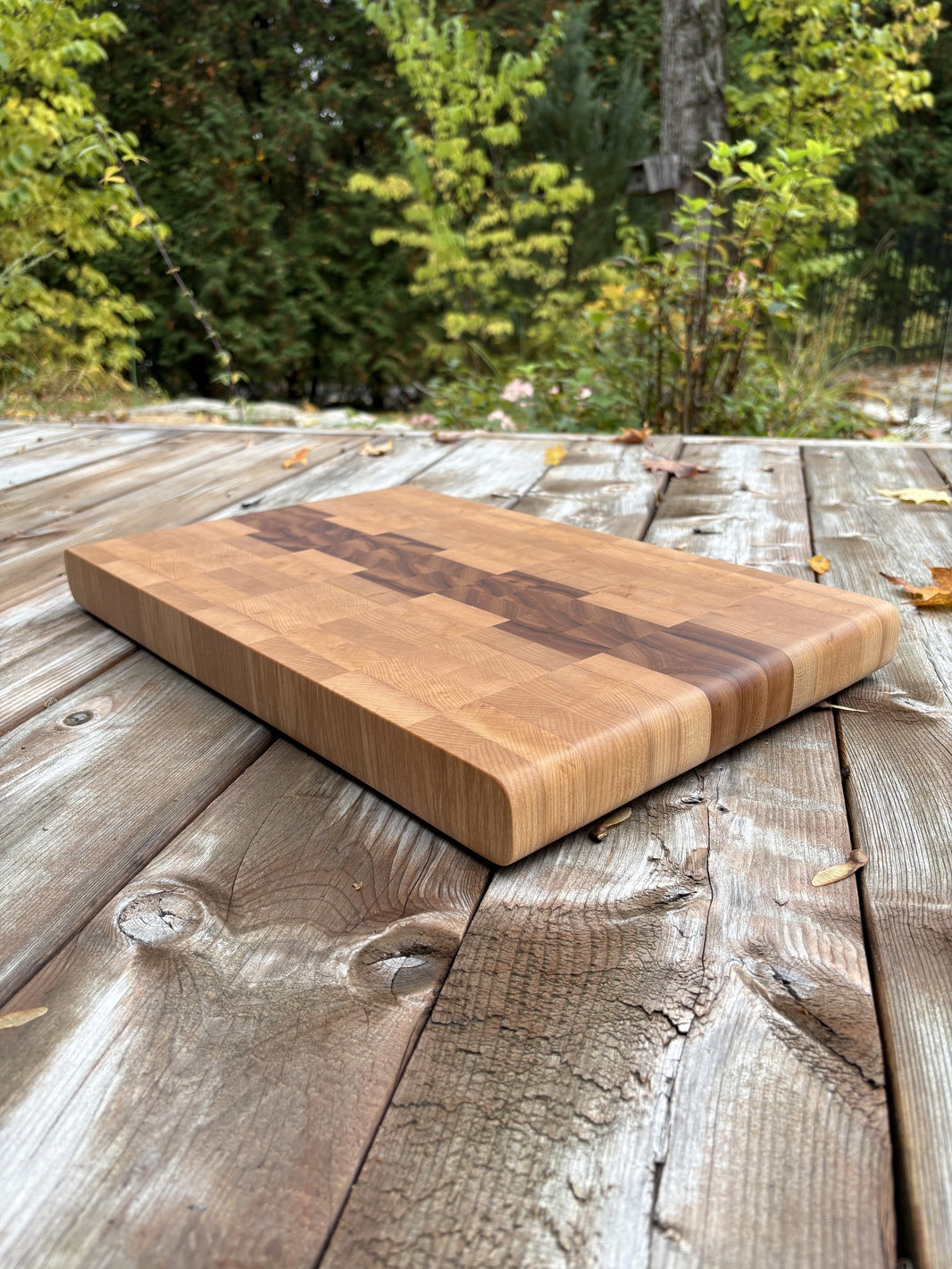 Planche à découper cerisier et érable en bois debout de 18,5 x 12,75 po, vue en angle sur une terrasse en bois, arbres en arrière-plan