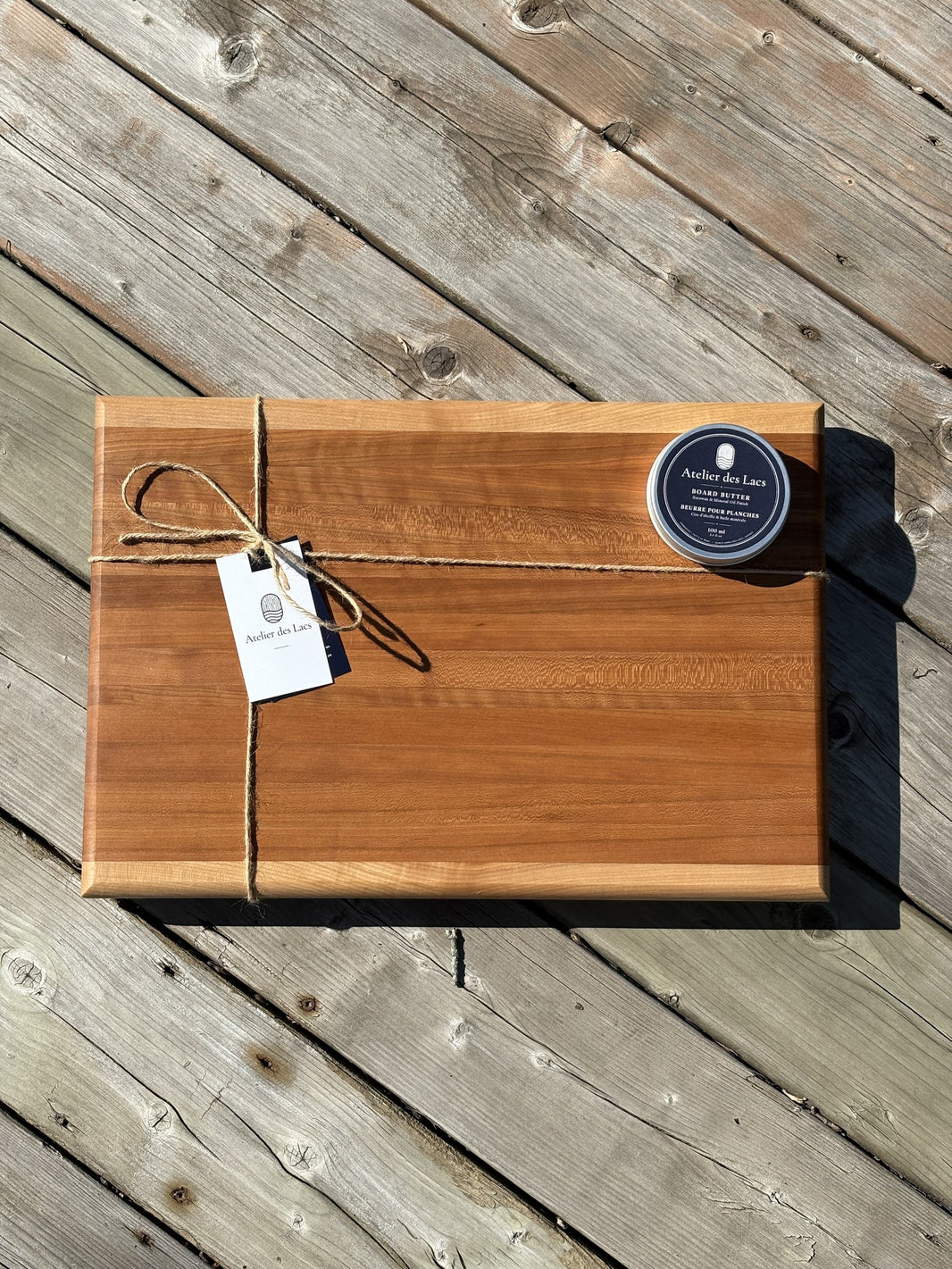 Cherry and maple cutting board 12 x 18 in (sold) tied with twine, with a tag and a board butter can, top view on a wood deck