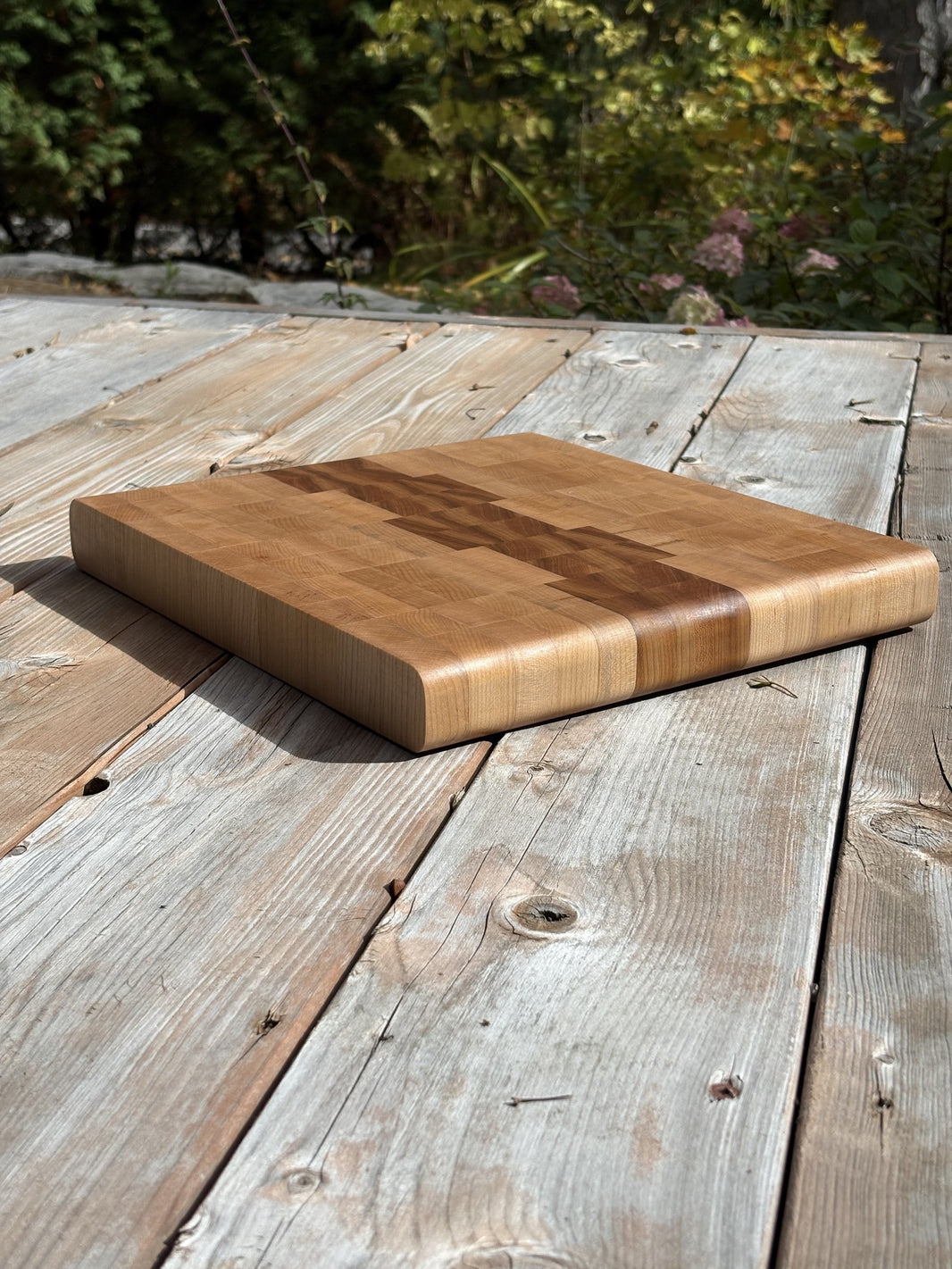 Planche à découper cerisier et érable en bois debout de 13 x 11,75 po, vue en angle sur une terrasse en bois, jardin en arrière-plan