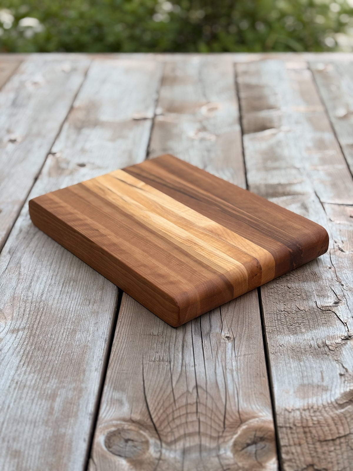 Petite planche à découper cerisier et érable de 11,25 x 8,25 po (vendue) aux bords arrondis, vue en angle sur une terrasse en bois